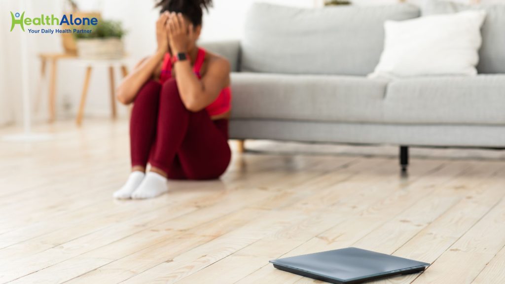 A person sits on a light wooden floor in a living room, hands over their face, with a tablet nearby and a gray sofa in the background; HealthAlone logo in the top-left corner.